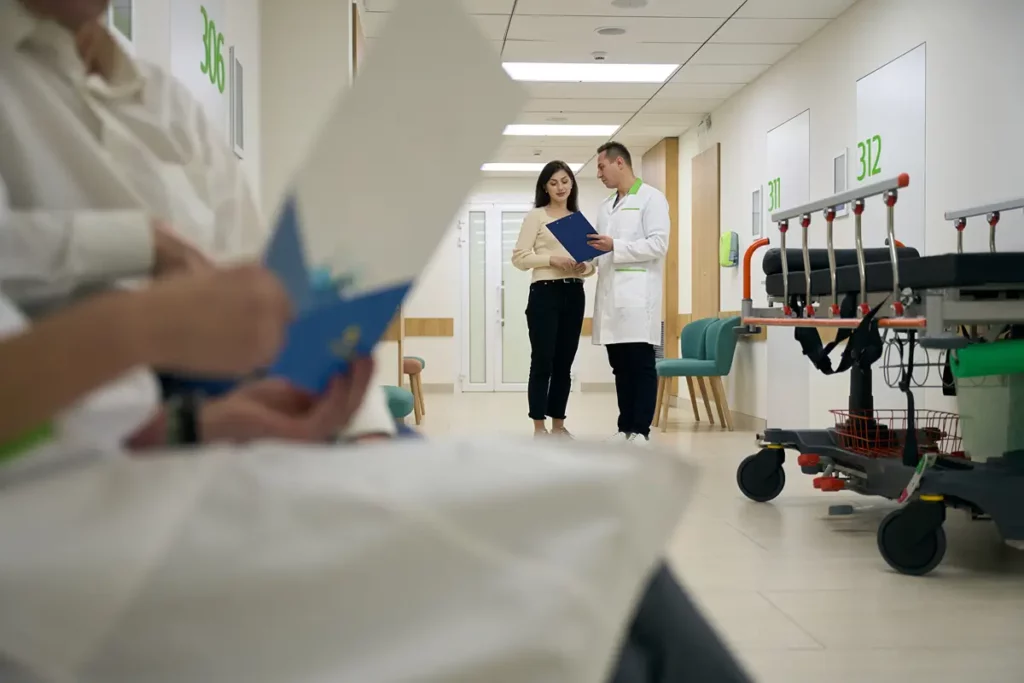 A doctor in a white lab coat is seen talking to a female patient in a hospital hallway, holding and looking at documents. Further in the foreground, a person in medical attire is blurred, holding a blue folder, partially obscuring the view. Hospital beds or gurneys are visible along the hallway, reinforcing the medical setting. The image suggests a serious discussion about medical care or administrative procedures.