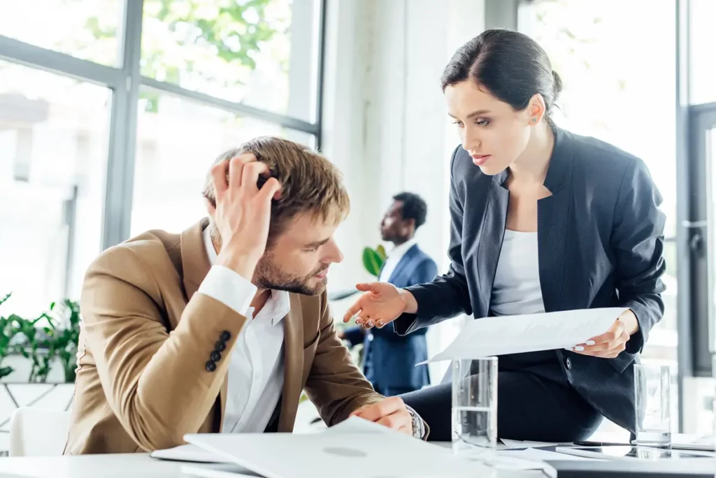A man in a brown suit and white shirt sits at a desk, looking stressed with his hand on his head, while a woman in a dark suit leans over, holding documents and speaking intently to him. Her expression is serious, and she appears to be explaining something important. In the background, another person in a suit is blurred, suggesting an office environment. The image conveys a high-stakes discussion, possibly involving difficult news or problem-solving.