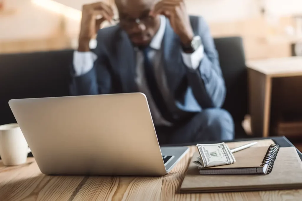 A man in a blue suit is seated in the background, out of focus, with his hands on his head, conveying stress or frustration. In the sharp foreground, a desk holds a laptop, a notebook with a pen, a stack of dollar bills, and a white mug. The image suggests financial worries, work-related stress, or difficult decisions, possibly related to bankruptcy or taxes.
