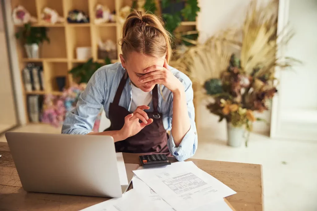A young woman with her hair tied up is seated at a wooden desk, covering her face with one hand, appearing stressed or overwhelmed. She is wearing a blue shirt and a brown apron. A laptop, a calculator, and scattered papers are on the desk in front of her. The background shows blurred shelves with various decorative items, suggesting a creative workspace or a small business setting. The image conveys financial stress or business difficulties.