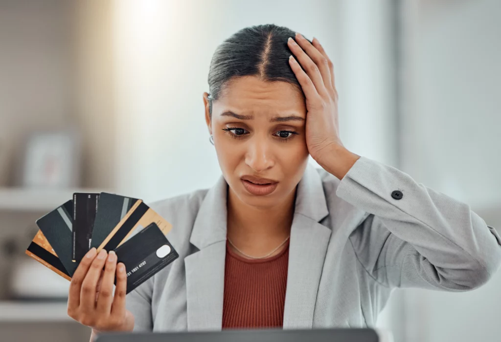 Woman in gray blazer holding multiple credit cards and looking worried, hand on head, illustrating credit card debt stress