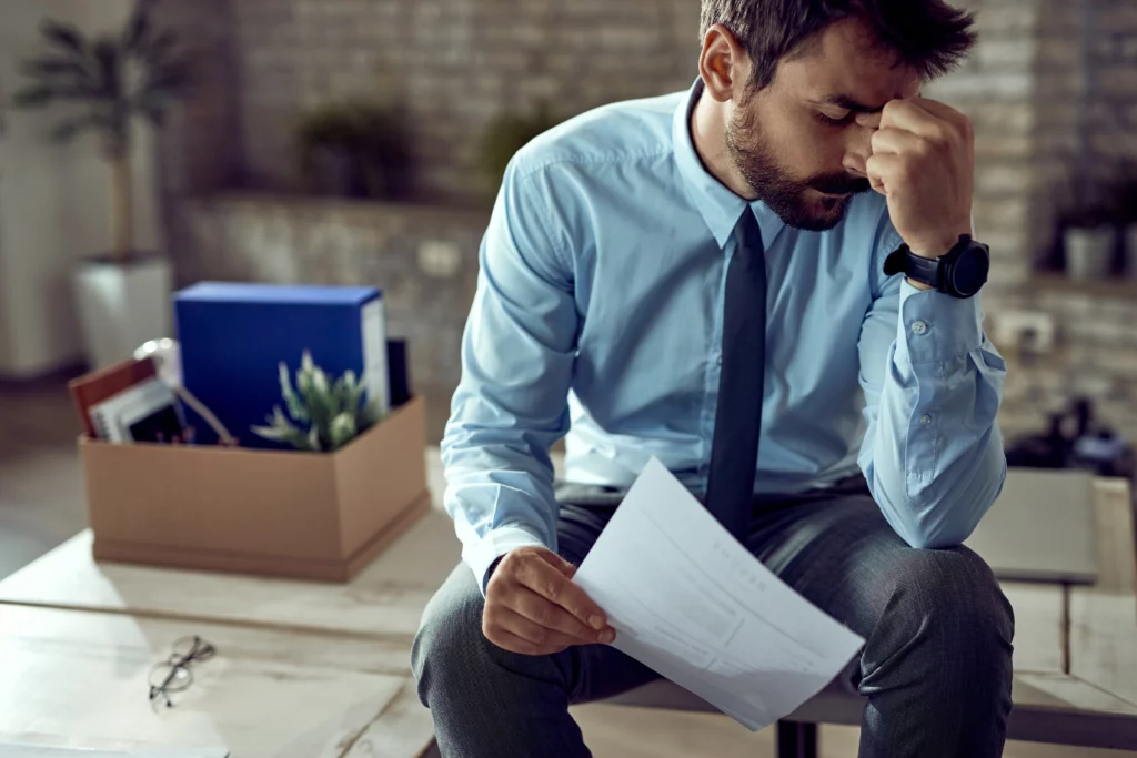 A distressed man with a beard, wearing a light blue shirt and dark tie, sits with his eyes closed and a hand on his forehead, holding a document. Next to him on a table, a cardboard box filled with personal belongings, including a blue binder and a plant, signifies a recent job loss or departure. A pair of glasses is also on the table. The image conveys feelings of sadness, worry, or a difficult transition.